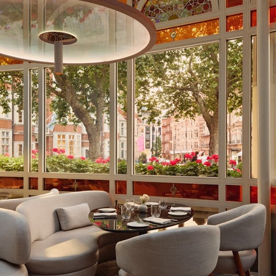 Intimate corner at Jean-Georges at The Connaught, with pale curved seating, a round black table set for four, and views of leafy Mayfair framed by red stained-glass panels and arched windows.