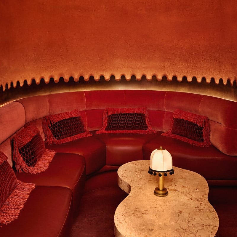 Curved banquette seating in rich red velvet at The Red Room, accented with fringed cushions and a sculptural marble table under ambient lighting.