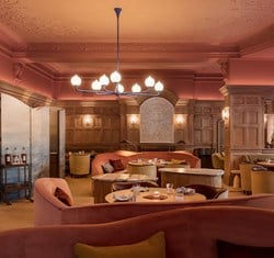 Warmly lit restaurant interior with blush pink ceiling, wood-panelled walls, and curved velvet banquettes, arranged beneath a sculptural chandelier and soft ambient lighting.